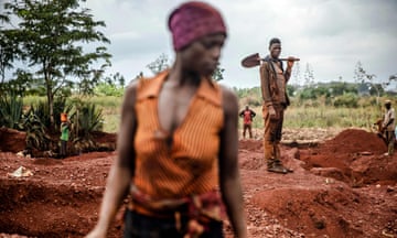 A male gold miner holding a spade in the background observes a female gold miner in an open-pit gold mine