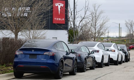 A line of Tesla cars sits outside of a building with the red Tesla logo on it.