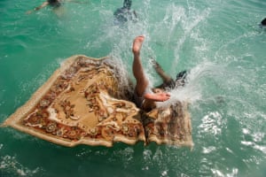 A brown-skinned man lands on his back on a Persian rug that is floating in water. The splash hides his face