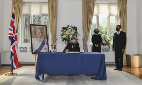 Wang Qishan sat on a table signing the condolence book next to a UK flag and a large picture of the Queen on an easle. Caroline Wilson and another staff member stand next to him.