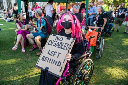 A young woman with bright pink hair, who wears a black facemask and is sitting in a wheelchair, holds a sign that reads: No disabled left behind.