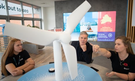 Keir Starmer, sitting at a table, listens as two young women explain something to him, pointing at a large model wind turbine on the table