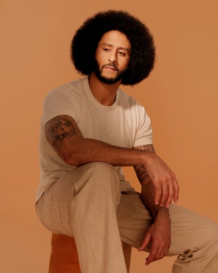 Colin Kaepernick sits on a stool with his hand resting on one leg; he is wearing a white T-shirt and smart-casual tan trousers and is shot against a light borw