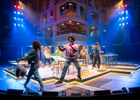 A cast dancing around an open set with a large tower structure in the middle and multicoloured lights above