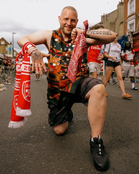 A bald man wearing shorts and a sleeveless top covered with Arsenal badges and a football scarf tied to each of this wrists kneels down in the road