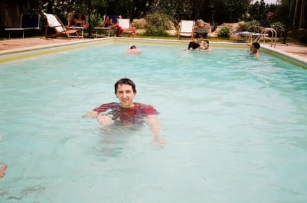 Stewart in a red T-shirt stands in ta fairly large swimmin pool looking at the camera, with people at the other end of the pool behind him.