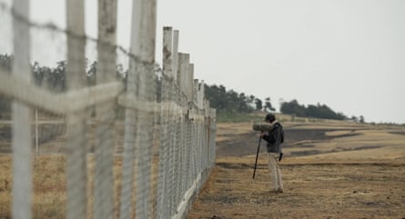 A man stands next to a wire fence in a barren landscape, holding a microphone.