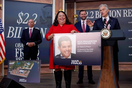woman in red dress smiles while holding a poster of a man smiling, as a man in suit next to her speaks at lectern with hand gestured