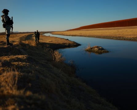 Two men in uniform patrol a sparse expanse of land. The border wall is visible in the distance