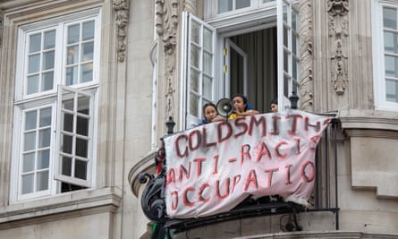 Two brown-skinned students, one with a megaphone, in 2019 on a balcony draped with a sheet with “Goldsmiths anti-racism occupation” written on it