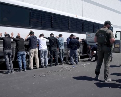 A row of man stand with their hands on a bus, as law enforcement officials talk behind them.