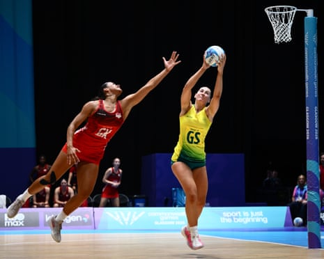 Two netball players jump for the ball, one in red reaching up, one in yellow catching it