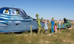In a grass field are a series of wood cut-outs painted to resemble a flying saucer, a tall green alien, and a white family next to a car with the hood up smiling at the alien.