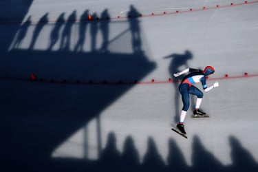 A speed skater leans forward racing on ice, with long shadows of spectators cast beside a red barrier