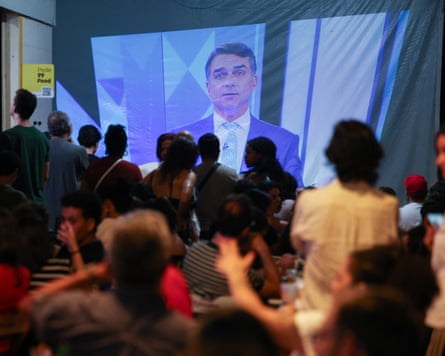 A crowd watches Flavio Bolsonaro speak on screen during a televised presidential debate.