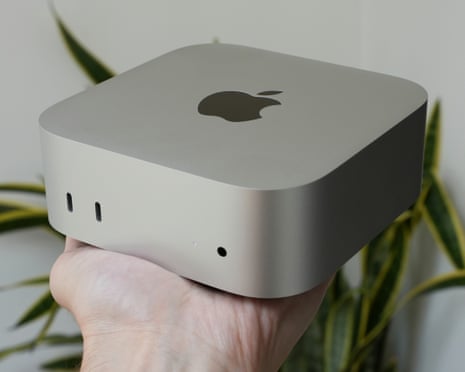 A hand holds up a small silver Mac mini M6 with an Apple logo on top.