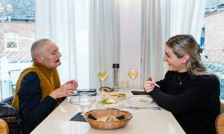 An older woman with very short cropped grey hair and bright red lipstick, wearing a mustard sleeveless polo neck over a black long-sleeve top, and a younger woman with long blonde hair pulled back from her face, wearing a black polo neck with a single pearl necklace, sitting chatting at a restaurant table