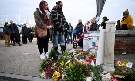 people attend a vigil outside Portland port: a man and woman pay their respects in front of a pile of flowers and a board of messages laid on the dockside. Others are seen standing quietly in the background.