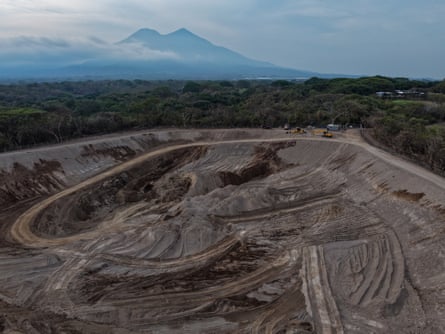 Aerial view of several hectares of land cleared in the middle of scrubby forest with heavy equipment parked at the edge. What looks like two volcanoes loom in the distance