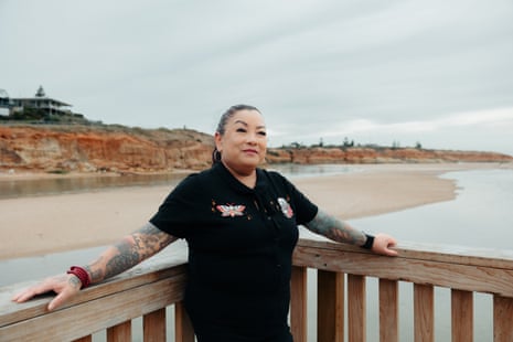 Chantal Doecke looking at the camera, standing with her arms outstretched on a wooden fence by the river, wearing a black T-shirt with two small logos on the front and black trousers