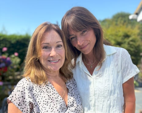 Davina McCall and Ann Forbes-Burford: Ann has shoulder-length blond hair and wears a white blouse with black spots; Davina is a little taller than her with darker hair and wears a white blouse. They are standing together outdoors in a garden with a hedge and blue sky behind them.