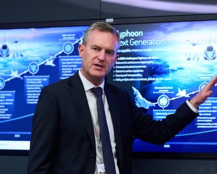 A man in suit gestures in front of a poster showing various images of fighter jets