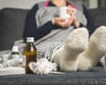 An unwell woman drinks a hot beverage with her legs up and medicine and scrunched-up hankies or tissues near her feet
