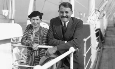 Ernest Hemingway, in a suit and tie, with a moustache and shiny hair parted on the right, leans on the rail of a ship next to his wife Pauline, in a suit dress and close-fitting hat, both slightly smiling into the sun