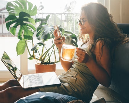 Young woman with laptop and phone sitting in living room and drinking tea.