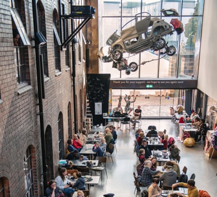 An atrium in a museum with people sitting at tables and a dismantled car hanging from the ceiling