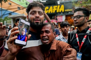 A man holding a phone with a picture of a woman puts his arm around the shoulder of another man who is crying