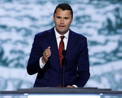 Charlie Kirk in a navy suit and red tie speaks at a podium with one hand raised.