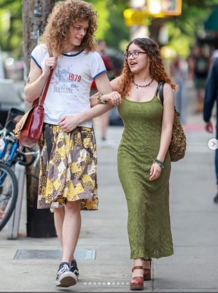 two people with curly hair walk with their arms linked. one is wearing a t-shirt and skirt and the other is wearing a long dress