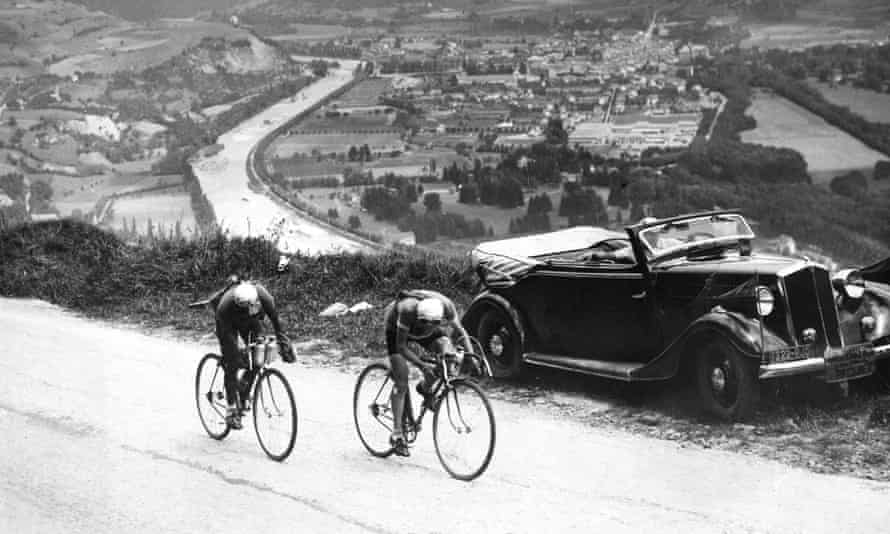 Two cyclists riding up a hill with 1930s open top car in background