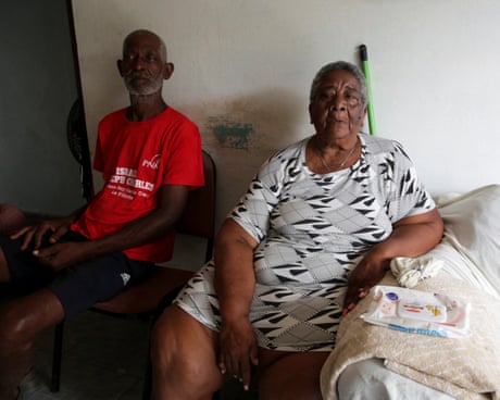 Two older people sit in a bedroom.