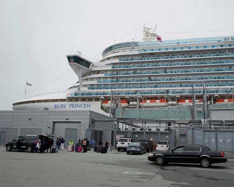A towering cruise ship docked at a cement dock with a few cars.