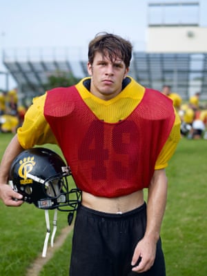 A young man wearing a gridiron uniform with big shoulder pads and holding his helmet under one arm and looking at the camera, on a football field