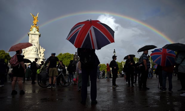 Londoners stand holding umbrellas outside near the Queen Victoria memorial opposite Buckingham palace under a grey sky with a rainbow. Several people are holding Union Jack-themed umbrellas