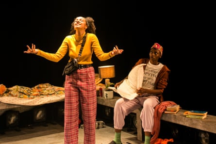 Stage shot showing two young woman in casual clothes, one standing with her arms stretched and looking upwards while smiling; the other sitting on a bench behind to the side her, looking at her with concern