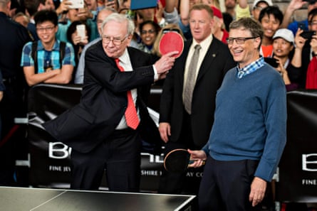 Two white men, one older and in a suit, the other younger and in a sweater and collared shirt, stand at one end of a ping-pong table.