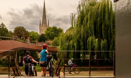 Group of cyclists take their bikes over a bridge across the river near the centre of Norwich. A cathedral spire is in the distance.