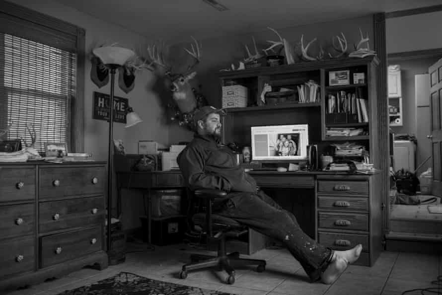 A man sits in an office chair next to a wood desk in his home office. Shelves are packed with papers and a deer head is mounted on the wall nearby.