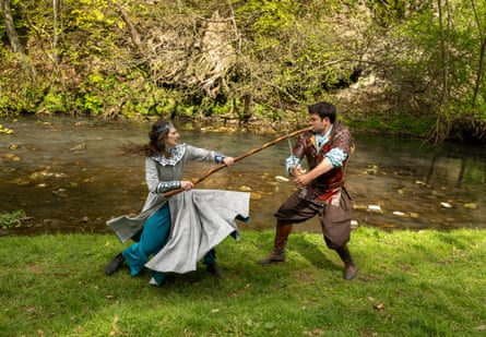 Two people dressed in medieval clothes battle with staff and sword in front of a river