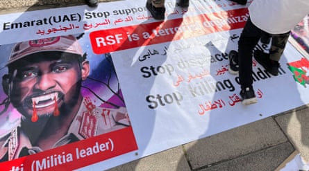 People walk on a poster showing a photo of an African man in military fatigues with cartoon-like fangs and blood over his mouth. There is writing in Arabic and English, saying “RSF is a terrorist organisation” and “stop killing children”