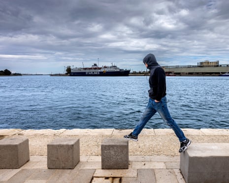 A young man wearing a dark hoodie, blue jeans and trainers jumping from one concrete block to another by the sea
