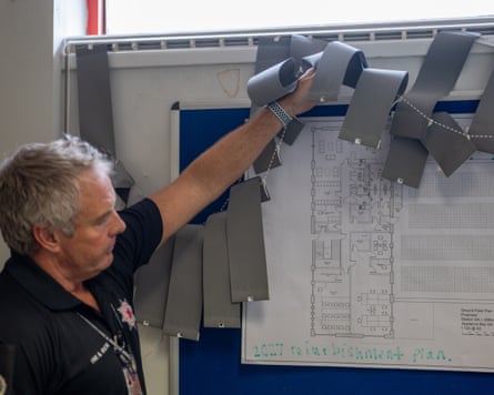 Carey holds up broken blinds to reveal a plan of the station stuck to a noticeboard. Written underneath it is ‘2027 refurbishment plan’