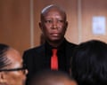 Julius Malema wearing a black shirt and jacket and red tie in court