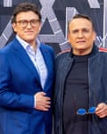 A man with rounded glasses and blue suit stands next to a shorter man with a jacket