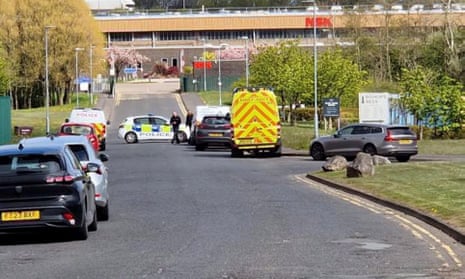 A police car blocks a road leading up to an industrial building, as an ambulance is also parked nearby