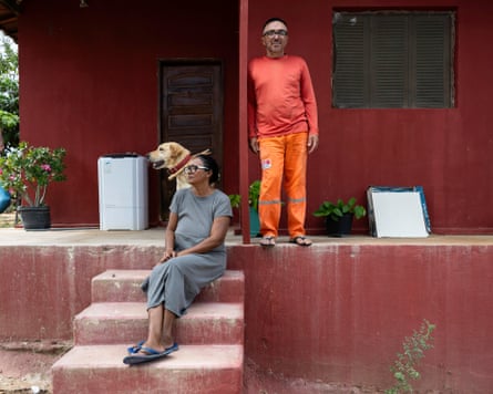 Two middle-aged people and a dog on the veranda of a house; a woman sitting on steps and a man standing, looking at the camera